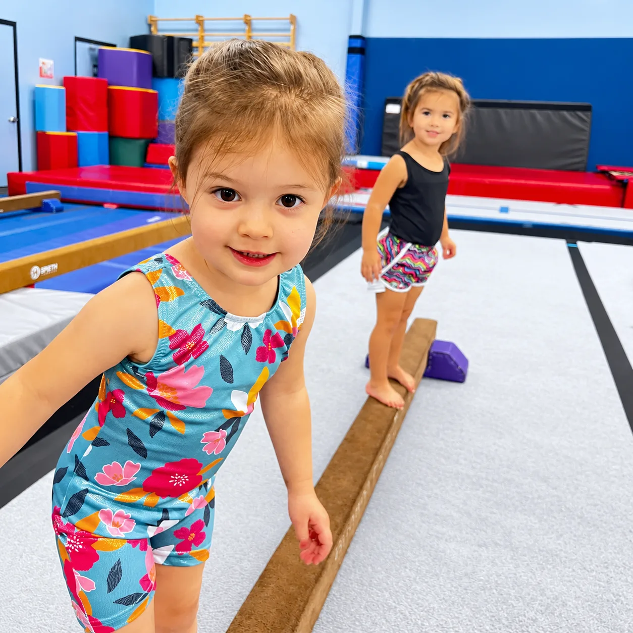 Two young gymnasts practicing on a low balance beam at All 4 Prosper.