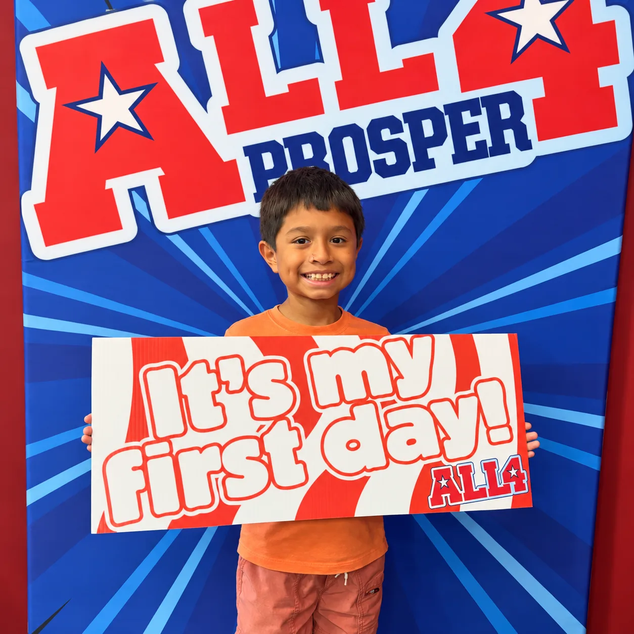 A smiling young athlete holding an “It’s my first day!” sign in front of the All 4 Prosper backdrop.