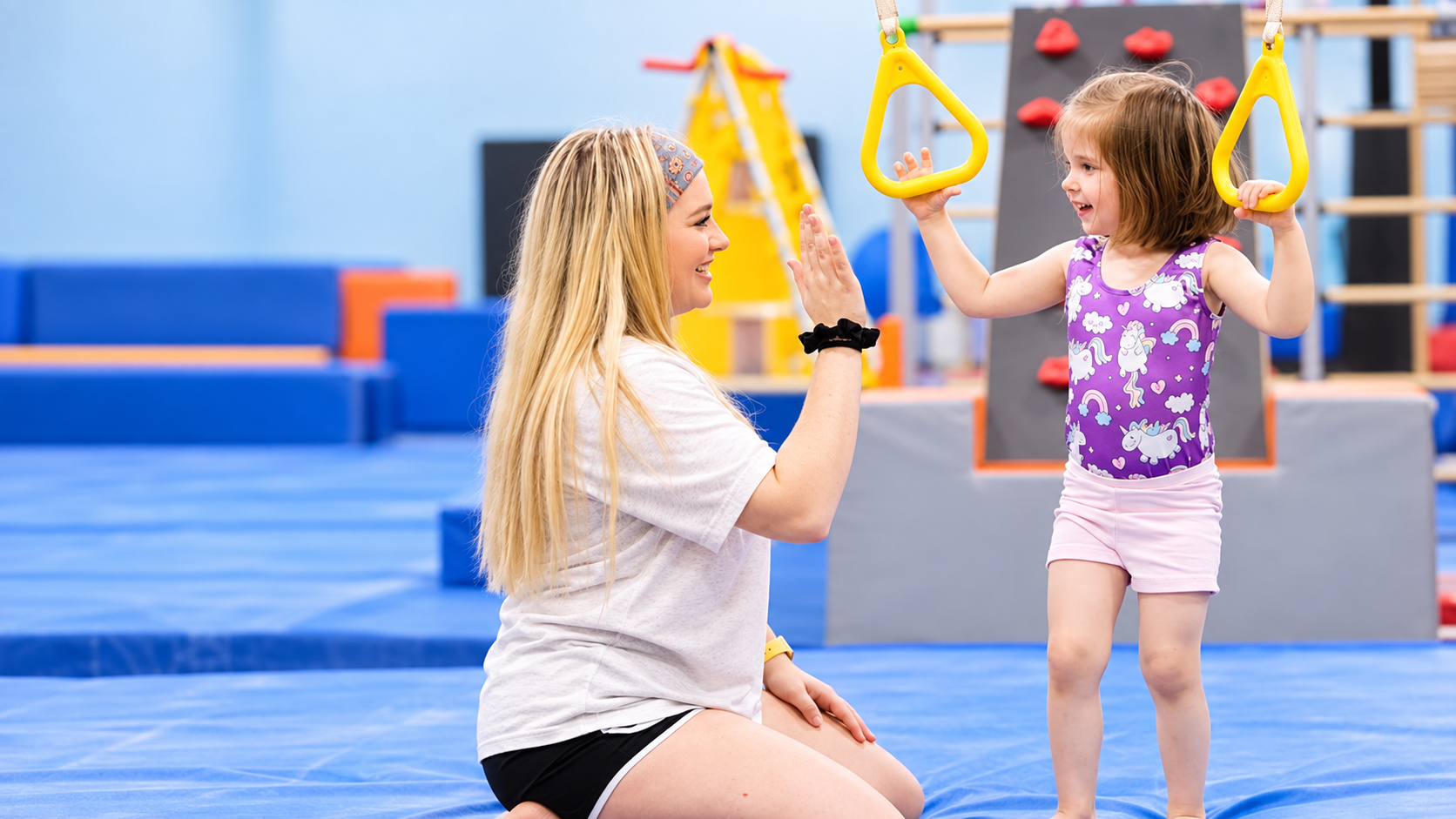 A coach kneels to high-five a smiling young gymnast holding yellow gymnastics rings.