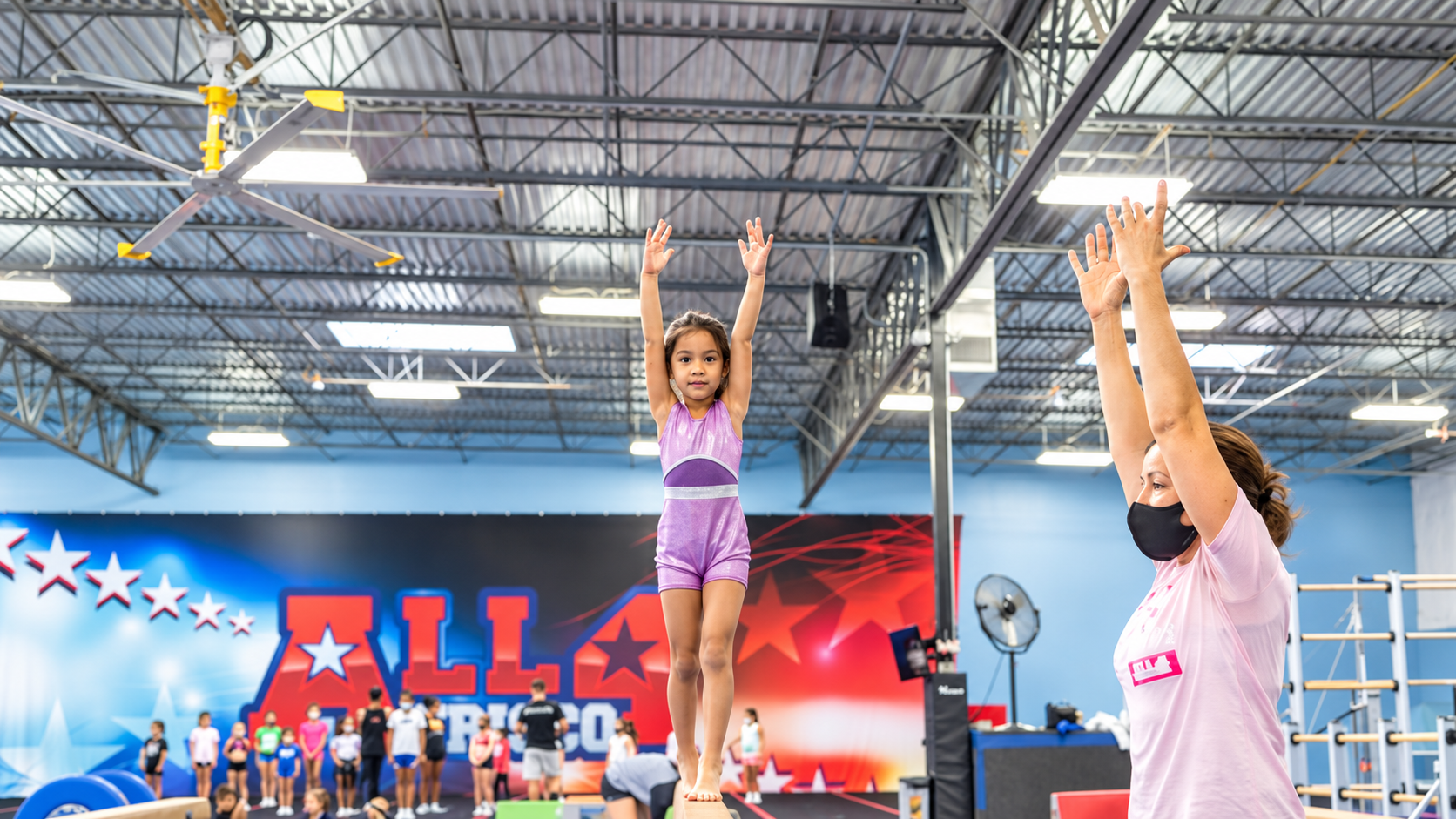 A young gymnast walks the balance beam with arms raised as a coach mirrors her pose